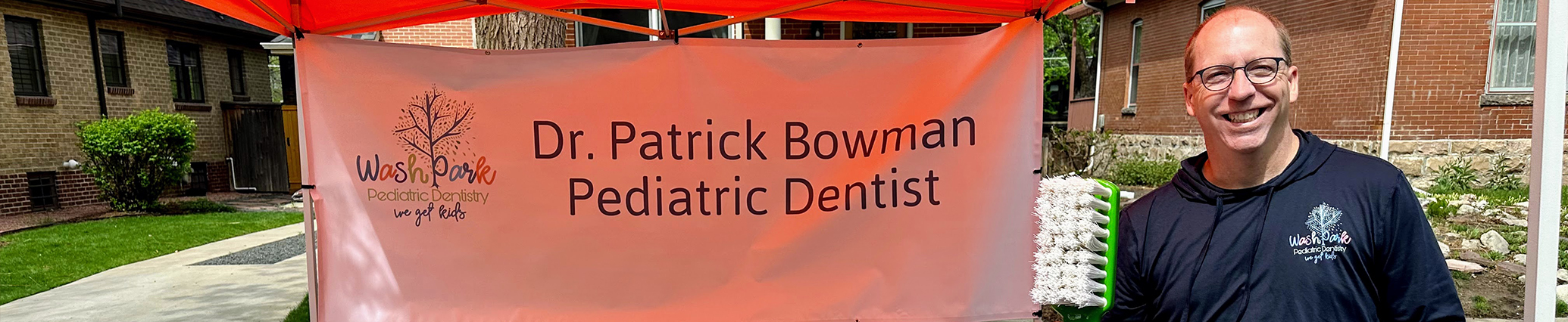 Doctor Patrick smiling with an outdoor poster that reads Doctor Patrick Bowman Pediatric Dentist