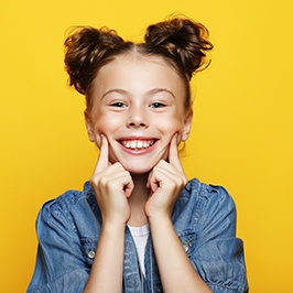 a little girl smiling and pointing to her cheeks in front of a yellow background