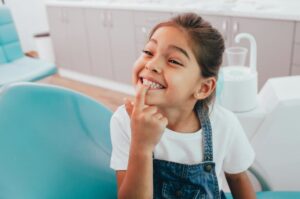 happy little girl smiling and pointing at her teeth in the dentist’s chair